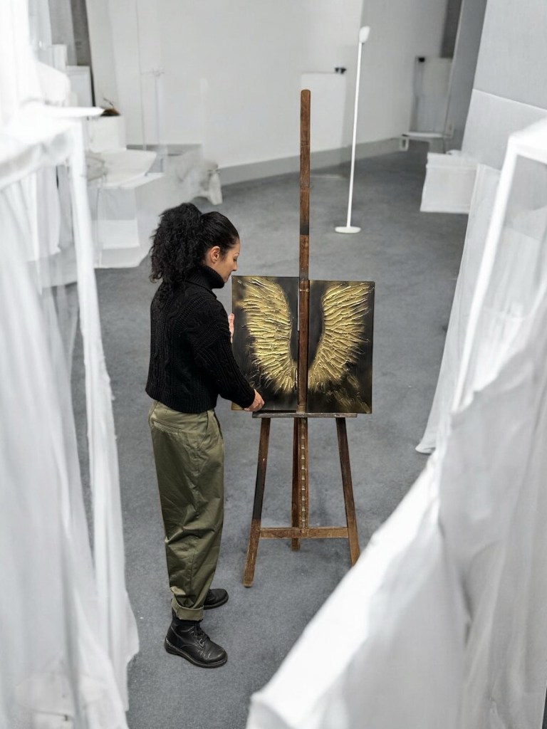 Claudia Ottone osserva un dipinto dorato a forma di ali esposto su cavalletto durante i preparativi di Medium Art Experience.